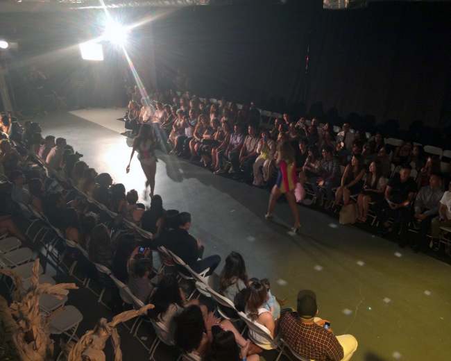 A fashion runway show is taking place with models walking down the catwalk in front of an audience seated on both sides.