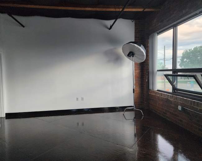 The image shows an empty room with a large window, exposed brick walls, and a photography light setup in the corner.