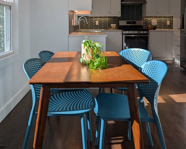 A wooden dining table surrounded by blue chairs sits in a bright kitchen with patterned backsplash and modern appliances.