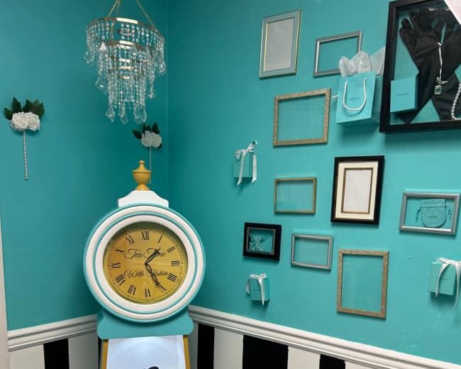 The image shows a turquoise wall with a vintage clock stand featuring a round clock, a small decorative black and white object, and framed pictures hanging nearby, along with a small chandelier.