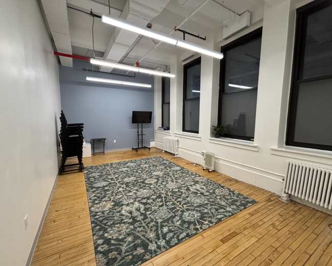 The image shows an empty room with a wooden floor, a large rug in the center, stacked chairs against one wall, and a small table with a television on the opposite side.