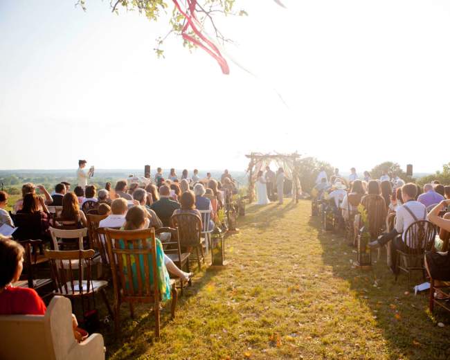 A wedding ceremony takes place outdoors on a grassy hill, with guests seated in various chairs facing an altar adorned with decorations.