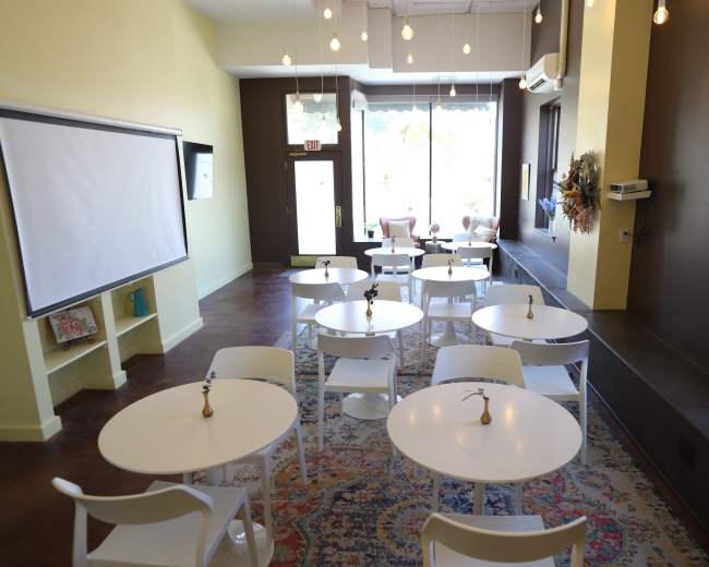 The image shows a bright, modern interior of a cozy café with several small white tables and chairs arranged on a patterned rug, along with large windows letting in natural light.