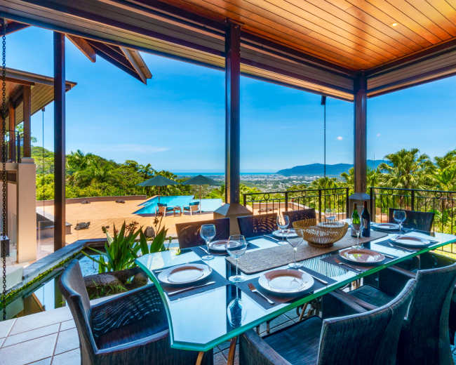 A spacious dining area features a glass table set for dinner, overlooking a scenic view of the mountains and ocean.