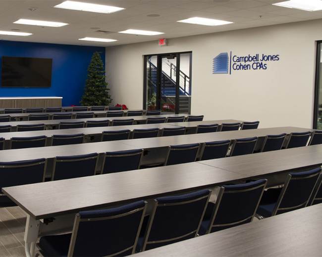 A modern, empty conference room with rows of tables and chairs facing two large screens on a blue accent wall.