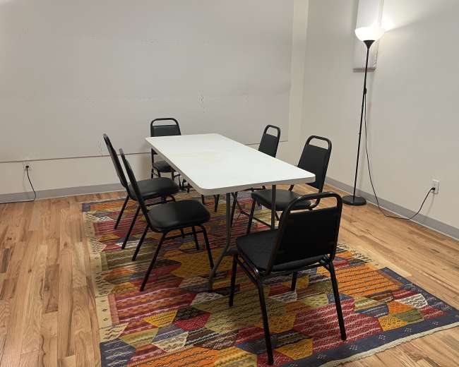 The image shows a simple room with a rectangular table surrounded by six black chairs on a colorful patterned rug, alongside a floor lamp in the corner.