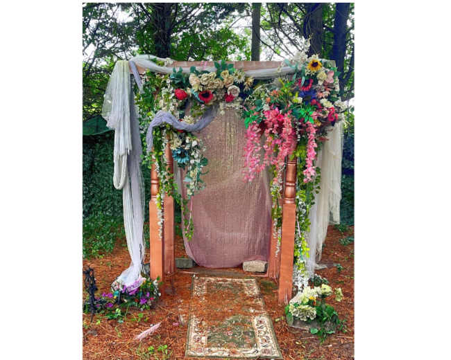 A floral-decorated archway with a draped fabric backdrop and a patterned rug on the ground in a natural setting.