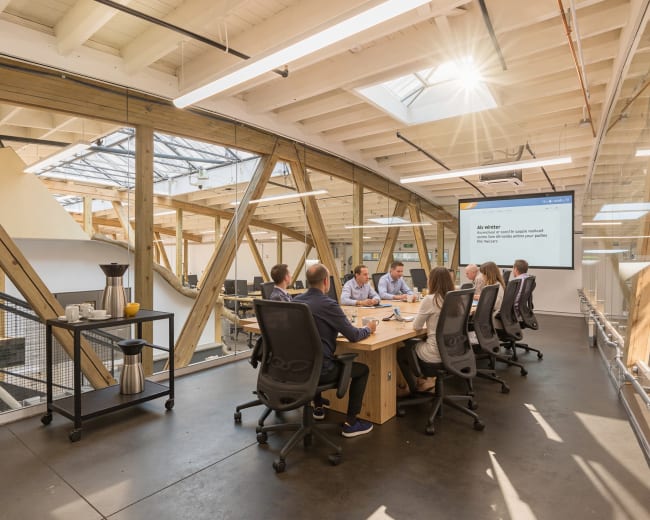A group of professionals is seated around a large wooden table in a modern conference room with large windows and a presentation screen.