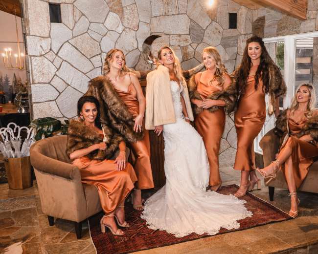 A bride in a white gown stands in the center with five women in satin dresses and fur wraps, all posing together in a stone-walled indoor setting.