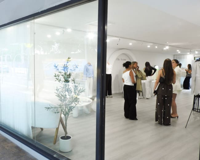 The image shows a modern boutique with large windows, featuring several people engaged in conversation and browsing clothing items inside.