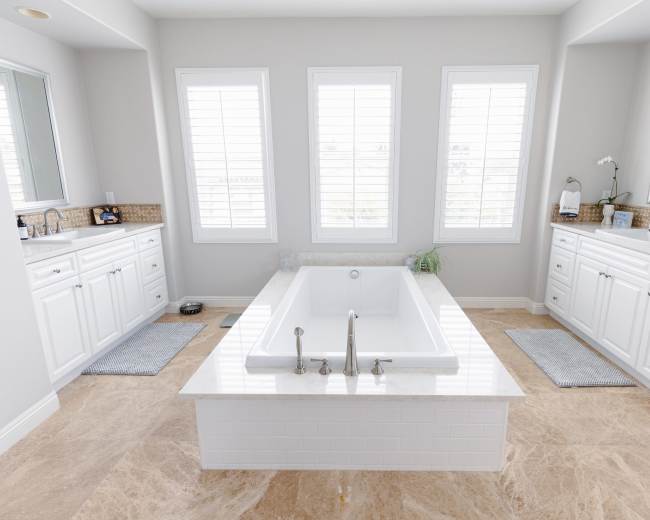 The image shows a spacious bathroom featuring a central freestanding bathtub, two vanities with sinks, and large windows with white shutters.