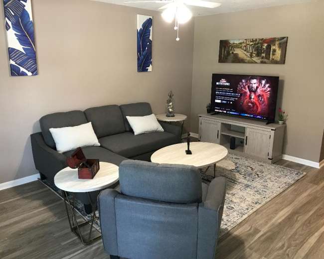 A living room with a gray sectional sofa, two small coffee tables, a television displaying content, and decorative wall art.