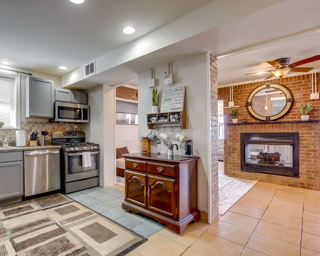 The image shows a modern kitchen with stainless steel appliances and a cozy living area featuring a brick fireplace and a round mirror.