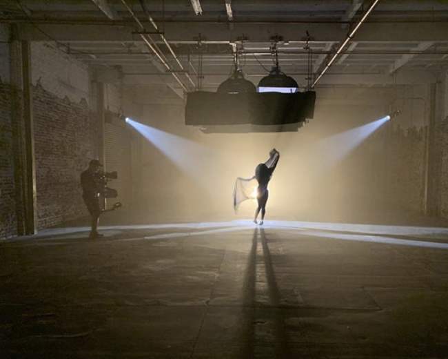 A silhouetted dancer poses dramatically in a dimly lit industrial space, while a camera operator captures the scene.