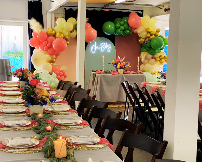 A long dining table is set with white plates and floral centerpieces, surrounded by colorful balloon decorations and a neon sign reading "On Baby."