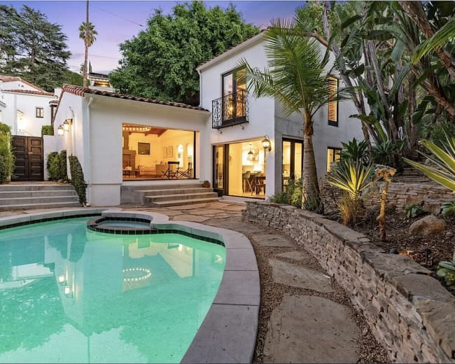 The image shows a private pool surrounded by stone pathways and tropical plants, with a two-story house featuring large windows and an open living area visible in the background.