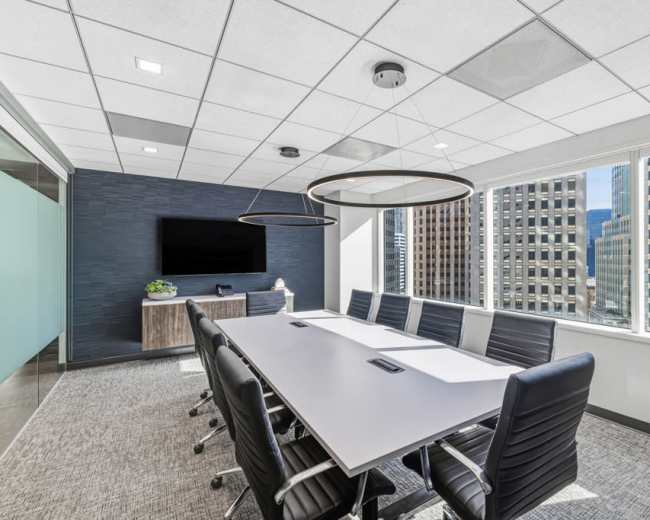 A modern conference room features a long table with ergonomic chairs, large windows offering a city view, and a wall-mounted screen.