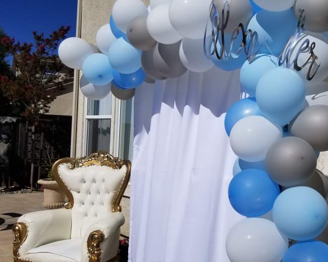 A balloon arch in shades of blue, gray, and white, with a gold-accented chair positioned nearby.