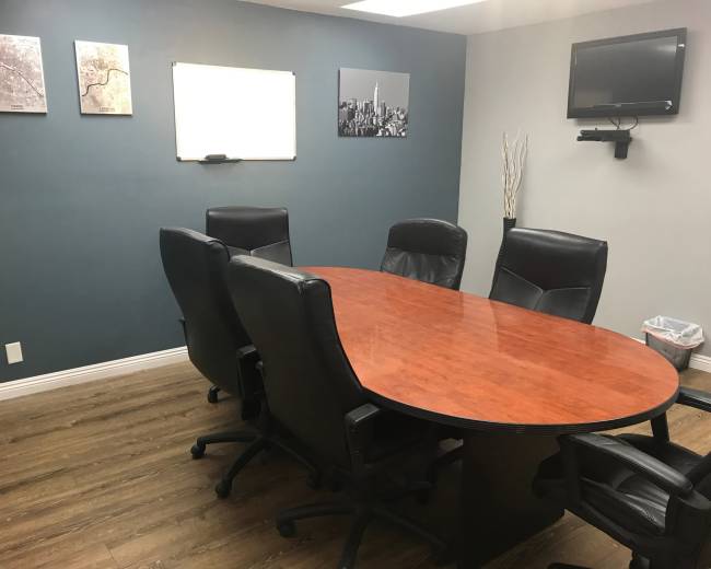A conference room features a wooden oval table surrounded by black leather chairs, with a whiteboard and wall-mounted TV on the gray walls.