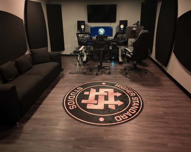 The image shows a modern music studio setup with a desk containing audio equipment, a large monitor, and a comfortable seating area with a black couch.