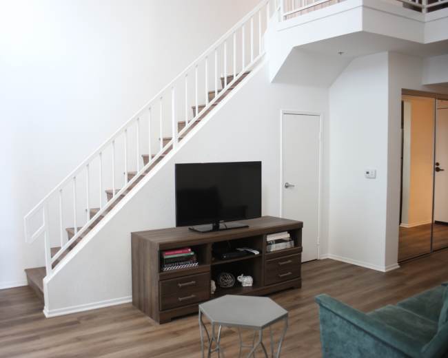 The image shows a living room with a television stand and a flat-screen TV positioned against a wall near a staircase leading to an upper level.