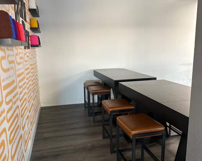The image shows a modern interior space featuring a long black table with four brown stools against a wall adorned with colorful craft supplies.