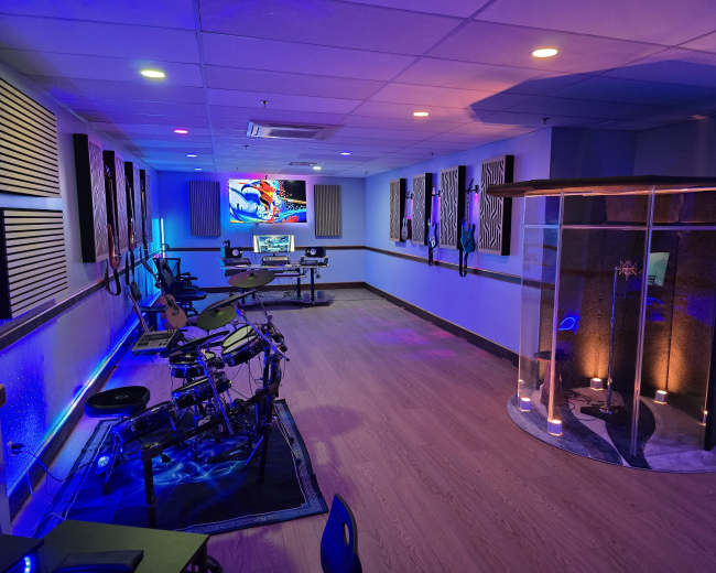 The image shows a brightly lit music room featuring electronic drums, guitars mounted on the walls, and a transparent structure illuminated with ambient lighting.