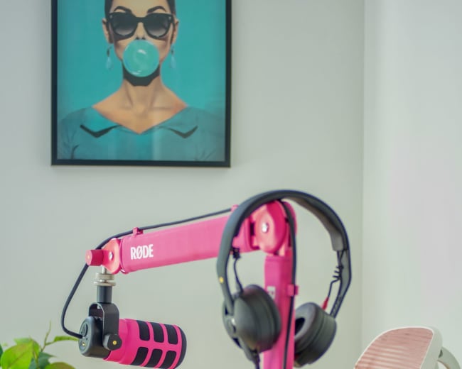 A pink microphone on a stand with headphones attached is positioned next to a laptop on a wooden table, beneath a framed portrait on the wall.