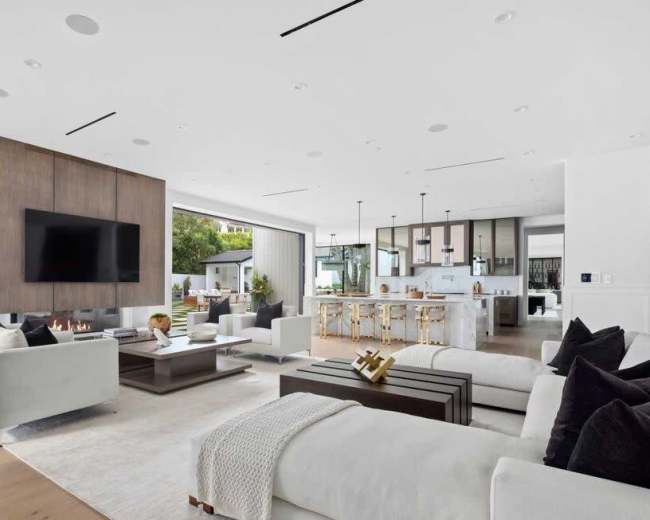 A modern living room features a large flat-screen TV mounted on a wooden panel, with white sofas arranged around a central coffee table and an open kitchen visible in the background.