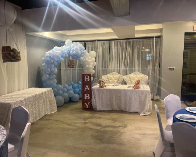 The scene features a decorated room for a baby shower, with white and blue balloon arch, two white chairs, a table draped in white cloth, and "BABY" spelled out in large letters.