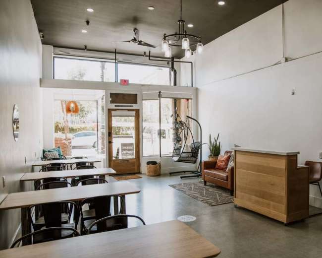 The image shows a bright, minimalist café interior with wooden tables, a reception area, a leather sofa, and large windows letting in natural light.