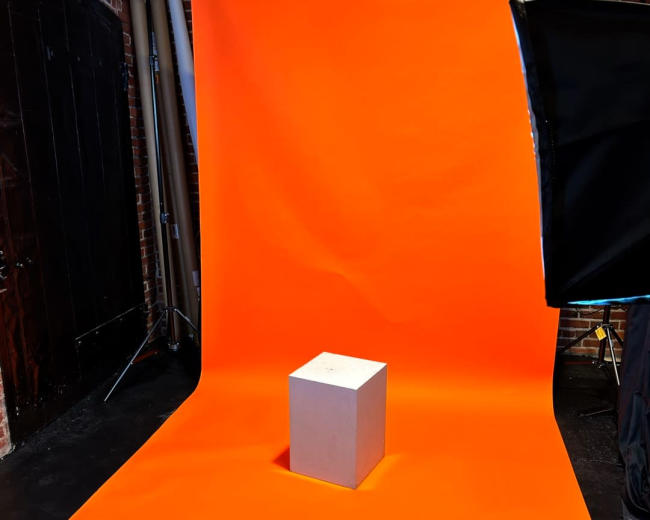 The image shows a gray cube placed on an orange backdrop in a studio setting.