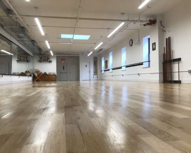 The image shows a spacious, well-lit dance studio with a wooden floor, mirrors, and a variety of exercise equipment against the wall.