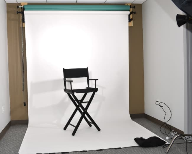 The image shows a simple photography setup featuring a black director's chair placed in front of a white backdrop in a studio.