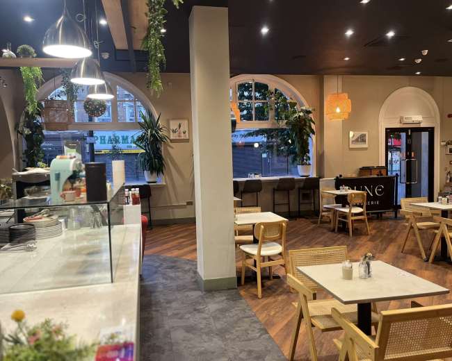 The image shows the interior of a café featuring wooden furnishings, large windows, and plants hanging from the ceiling.