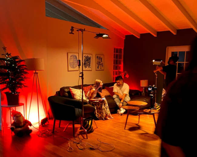 The scene depicts a cozy room with two individuals seated on a couch under warm red lighting, surrounded by various lighting equipment and a decorative tree.