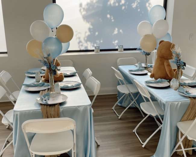 A decorated event space with light blue tablecloths, white folding chairs, and tables set for an occasion featuring balloons and a bear centerpiece.