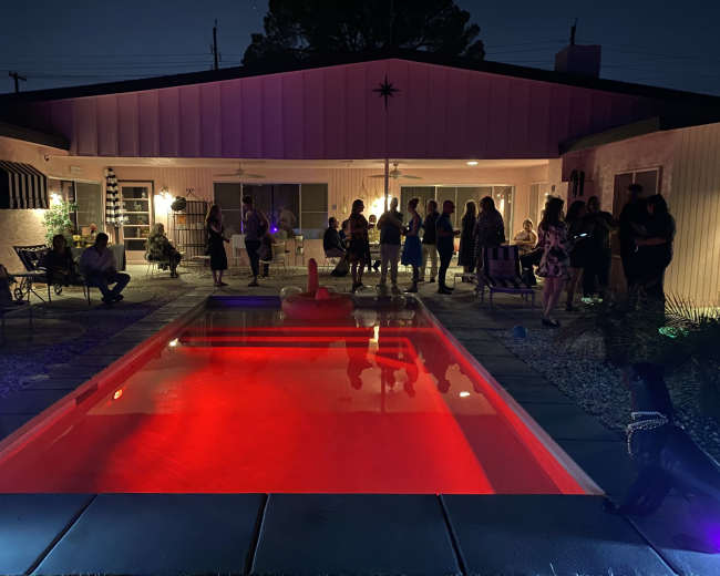 A group of people gathers around a brightly lit pool area in front of a house at night.