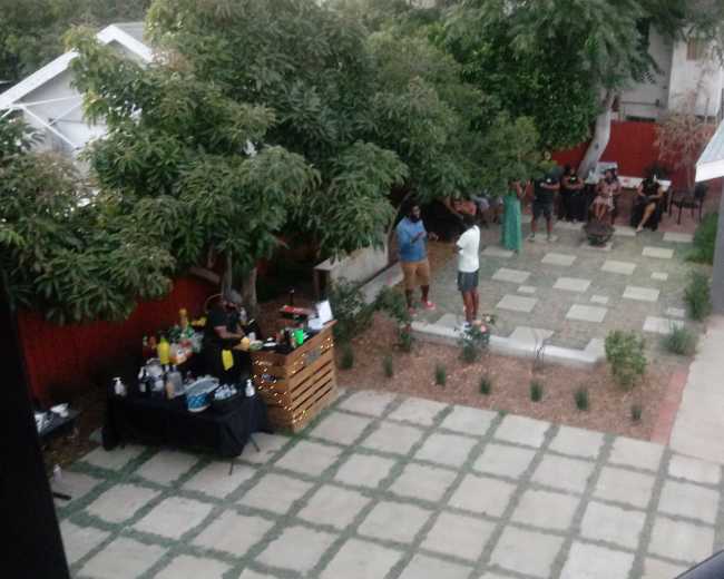 A group of people gathers in a backyard with a bar setup, seating, and greenery.