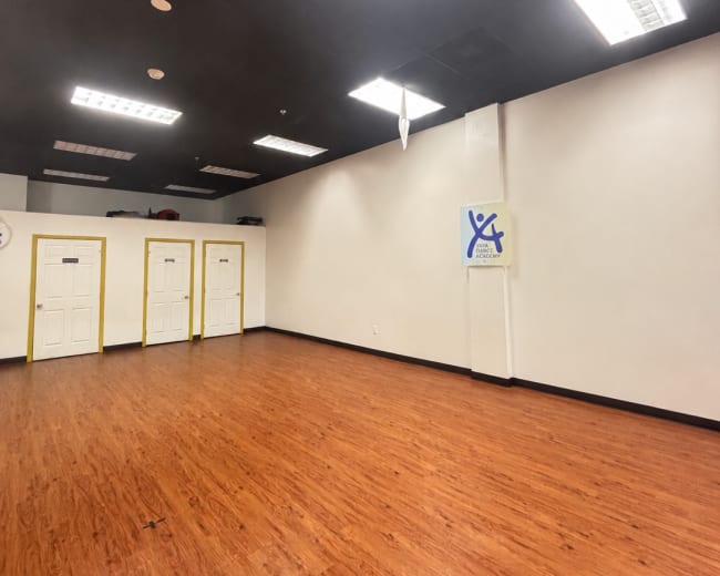 The image shows an empty room with wooden flooring, white walls, and three closed doors on one side.