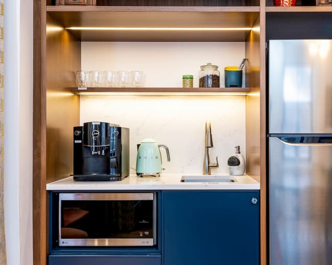 The image shows a modern kitchenette featuring a coffee maker, kettle, sink, and refrigerator, all arranged within wooden cabinetry with illuminated shelves.