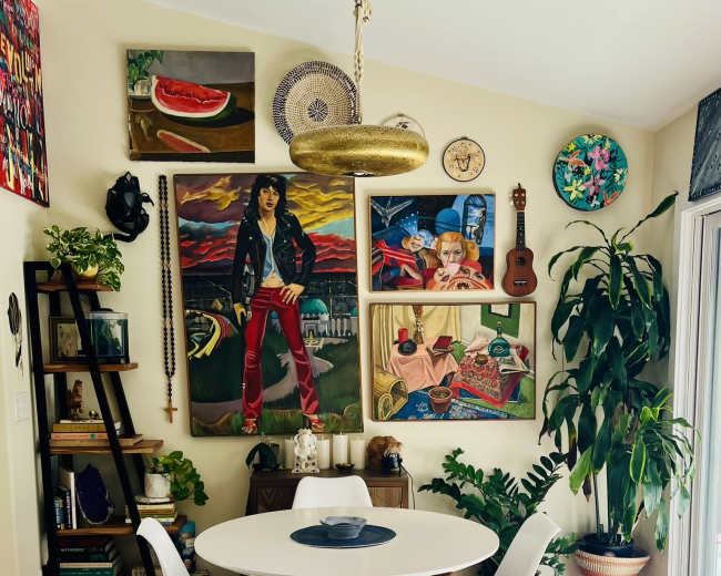 A dining area features a round table surrounded by white chairs, adorned with colorful wall art, plants, and decorative items.
