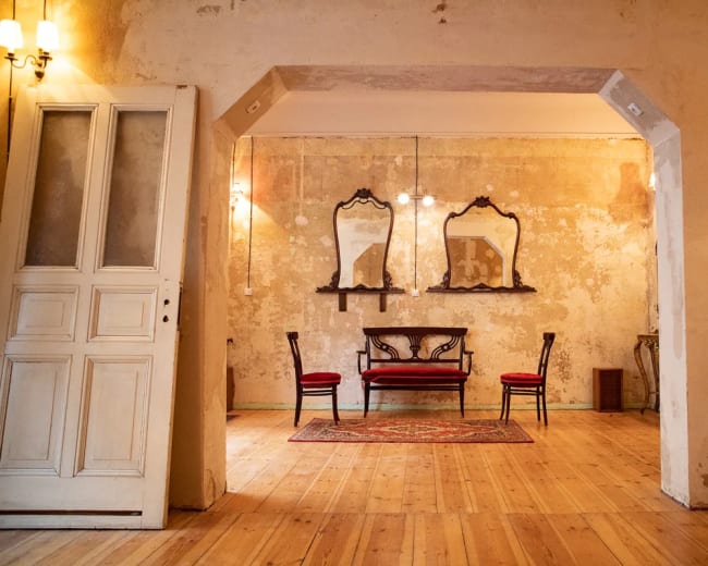 A room with a wooden floor, two red chairs, a central ornate bench, and three framed mirrors on the wall, all set against a peeling plaster backdrop.