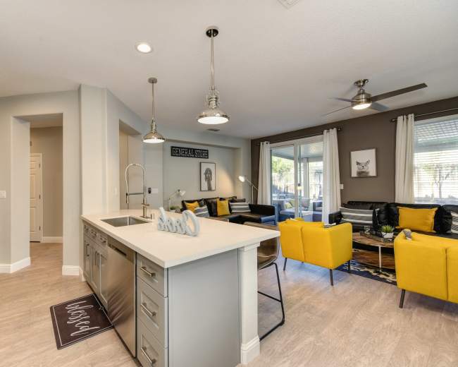 The image shows a modern open-concept living space featuring a kitchen with a central island, yellow accent chairs, and a cozy seating area with natural light.