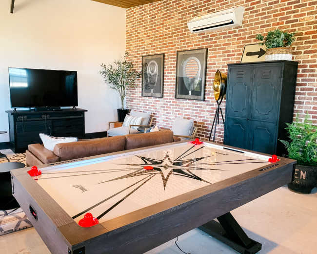 The image shows a game room with a long air hockey table, a brown sofa, a television, and decor featuring a brick wall and framed artwork.