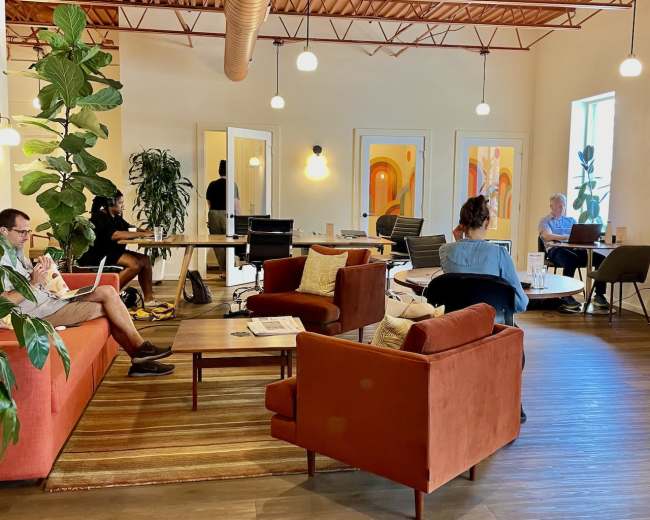 A shared workspace features several seating areas, plants, and individuals engaged in various activities like working on laptops and talking.