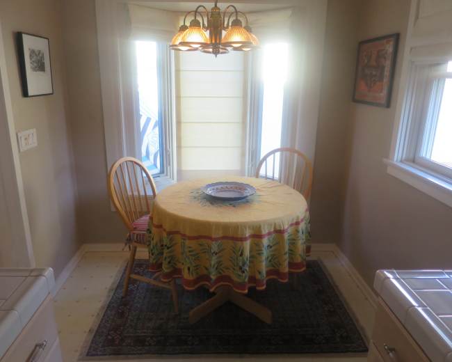 A small dining area features a round table with a yellow patterned tablecloth and two wooden chairs, surrounded by windows allowing natural light.