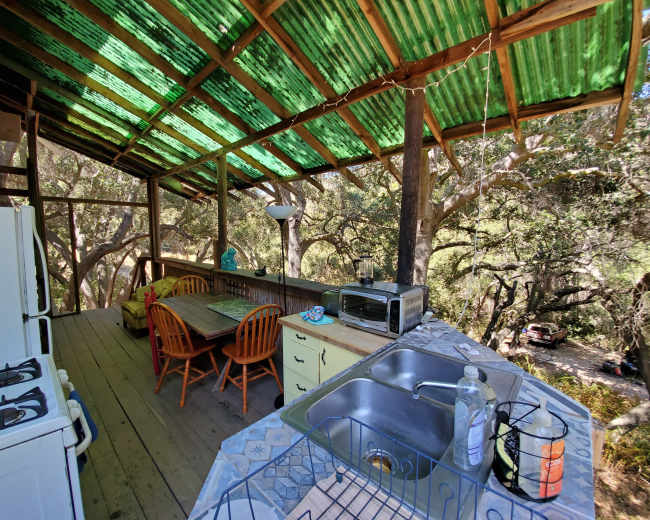 A rustic kitchen area with a wooden table and chairs, surrounded by trees, featuring a stove, sink, and a microwave under a green corrugated roof.