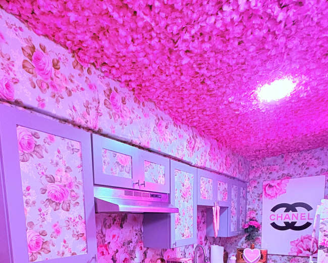 The image shows a kitchen with floral-patterned walls and ceiling, featuring pink decor, modern appliances, and a sign with the Chanel logo.