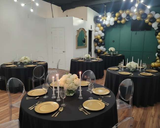 The image shows a decorated indoor event space with several round tables set for a formal gathering, featuring black tablecloths, gold plates, and floral centerpieces, alongside a balloon arch in black, gold, and white.